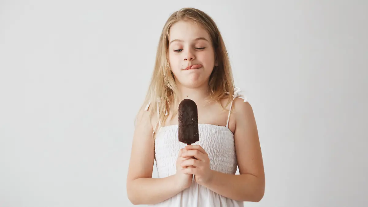 Close up of funny little girl with light long hair in white dress showing tongue, holding ice cream in hands, can't wait to eat it.