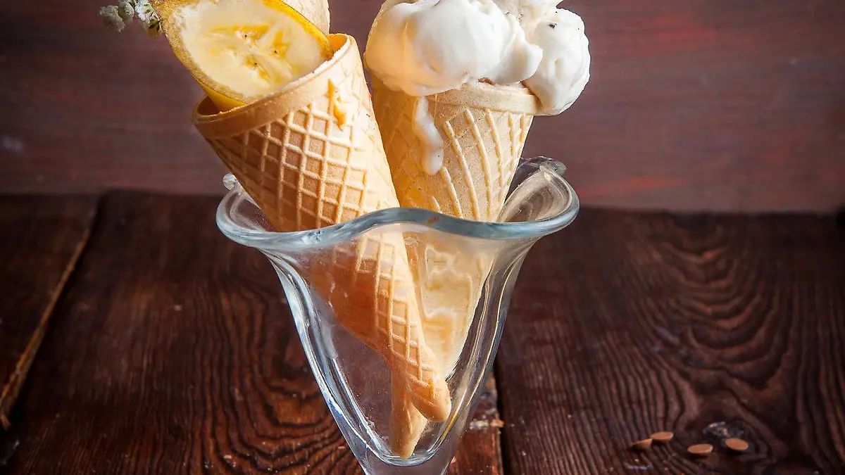 Side view waffle cones with banana ice cream and banana slices and chocolate drops in ice cream cup on wood background