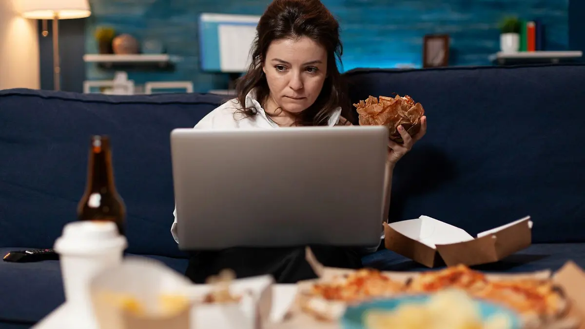 caucasian female sitting on sofa eating tasty delicious burger while working on laptop computer 123
