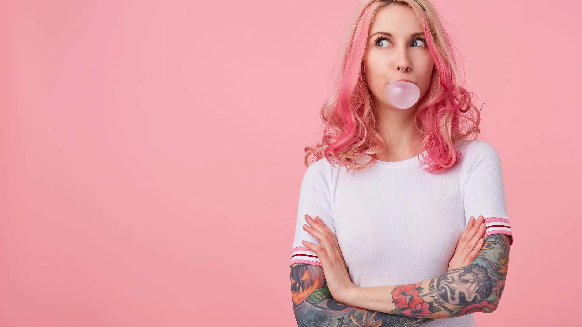 portrait-of-young-amazed-beautiful-pink-haired-lady-with-tattooed-hands-wears-in-white-t-shirt-blows-gum-ball-looking-to-the-left-in-surprise-stands-with-copy-space