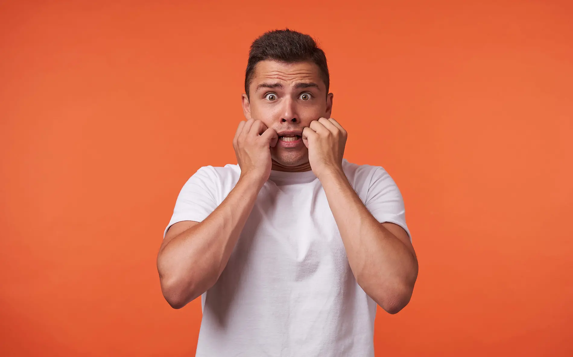 foto-estudio-joven-varon-pelo-bastante-castano-sosteniendo-manos-levantadas-mejillas-mientras-mira-miedo-camara-pie-contra-fondo-naranja